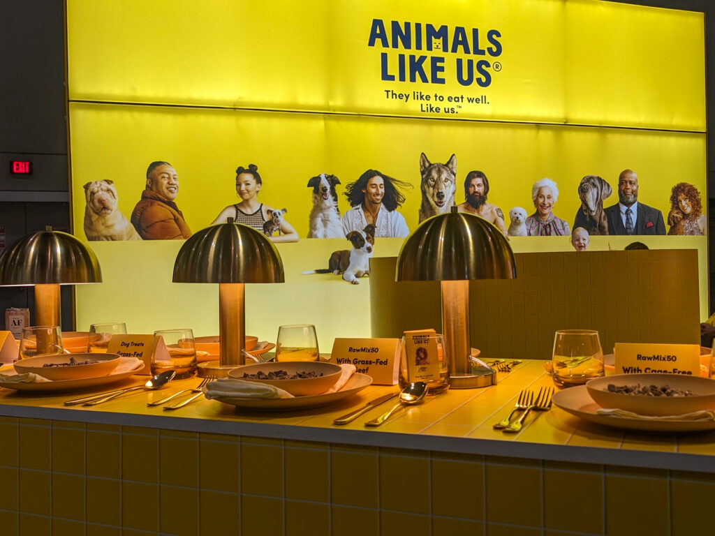 Animals Like Us booth in bright yellow and with a table set with fancy plates of freeze dried dog food at Global Pet Expo 2024
